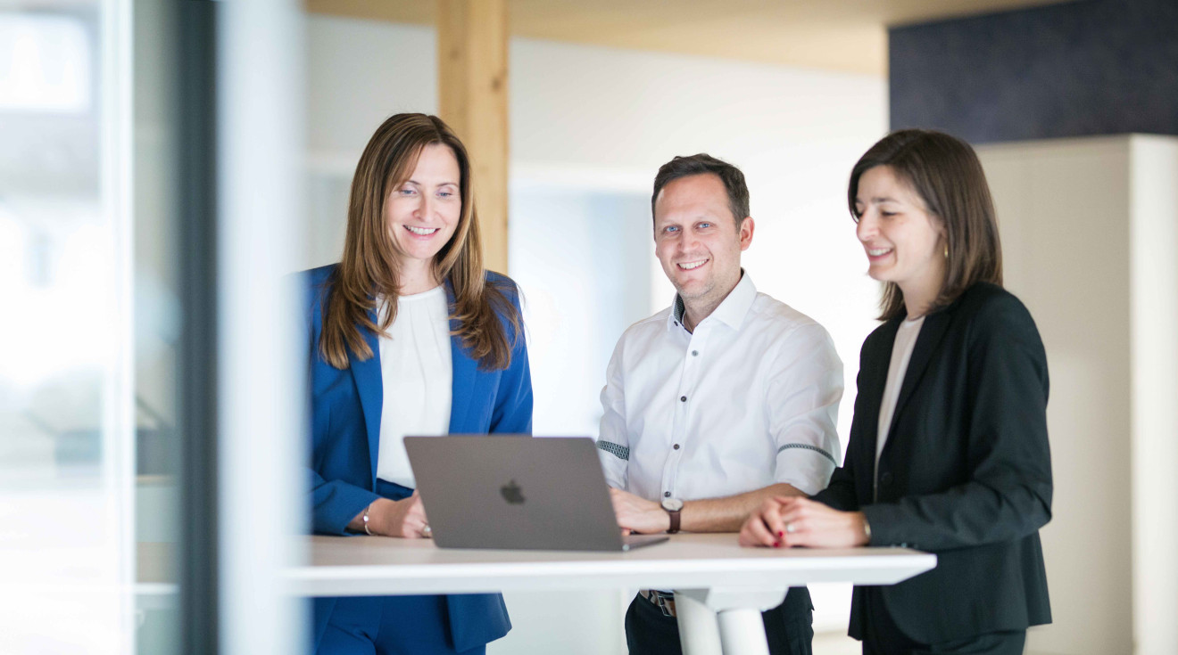 Zwei Frauen und ein Mann stehen im Büro und gucken auf einen Laptop